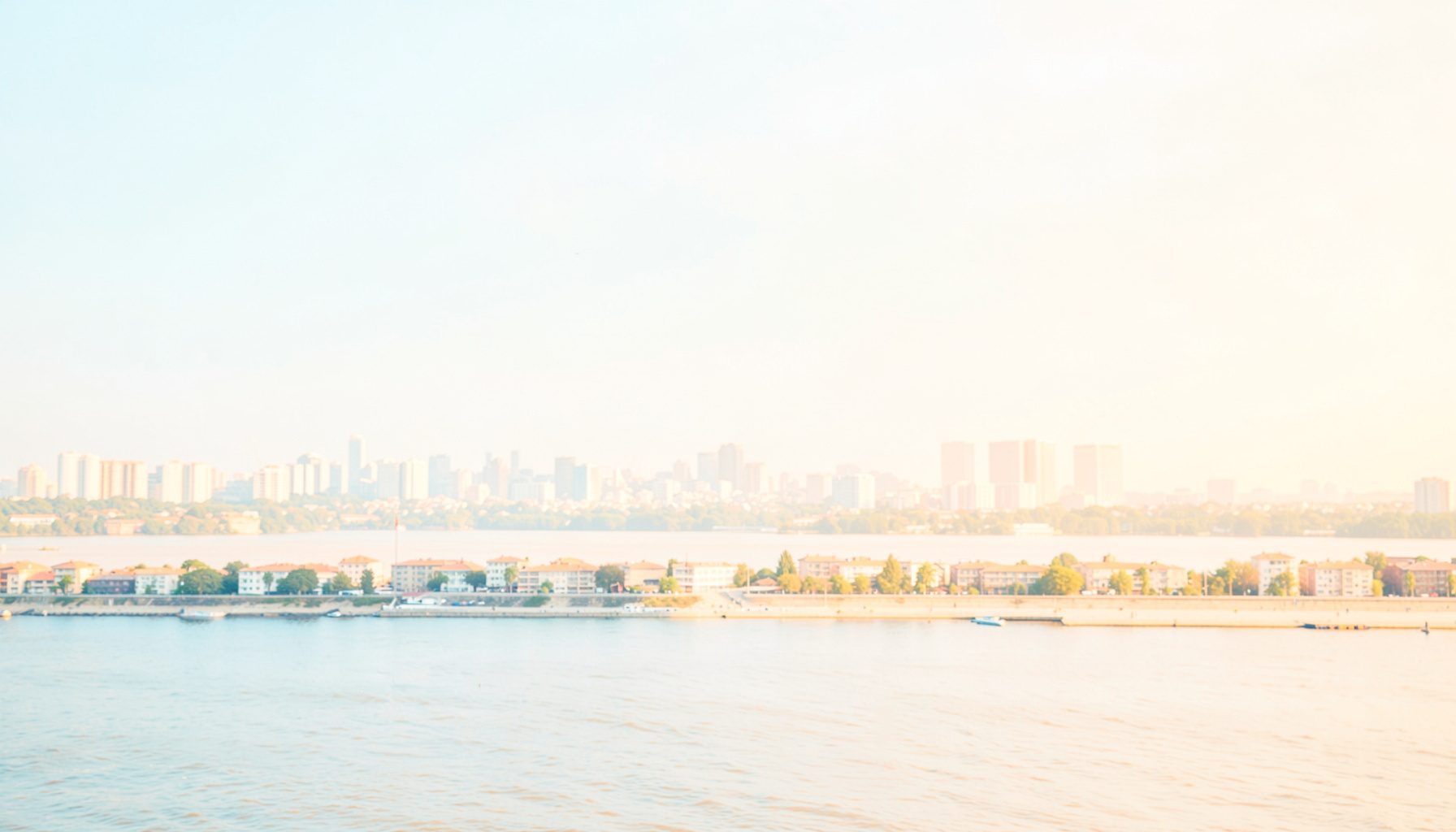 Bright, high-key photograph of a distant city skyline beside a wide river under a soft pale sky, in light blue and warm beige tones
