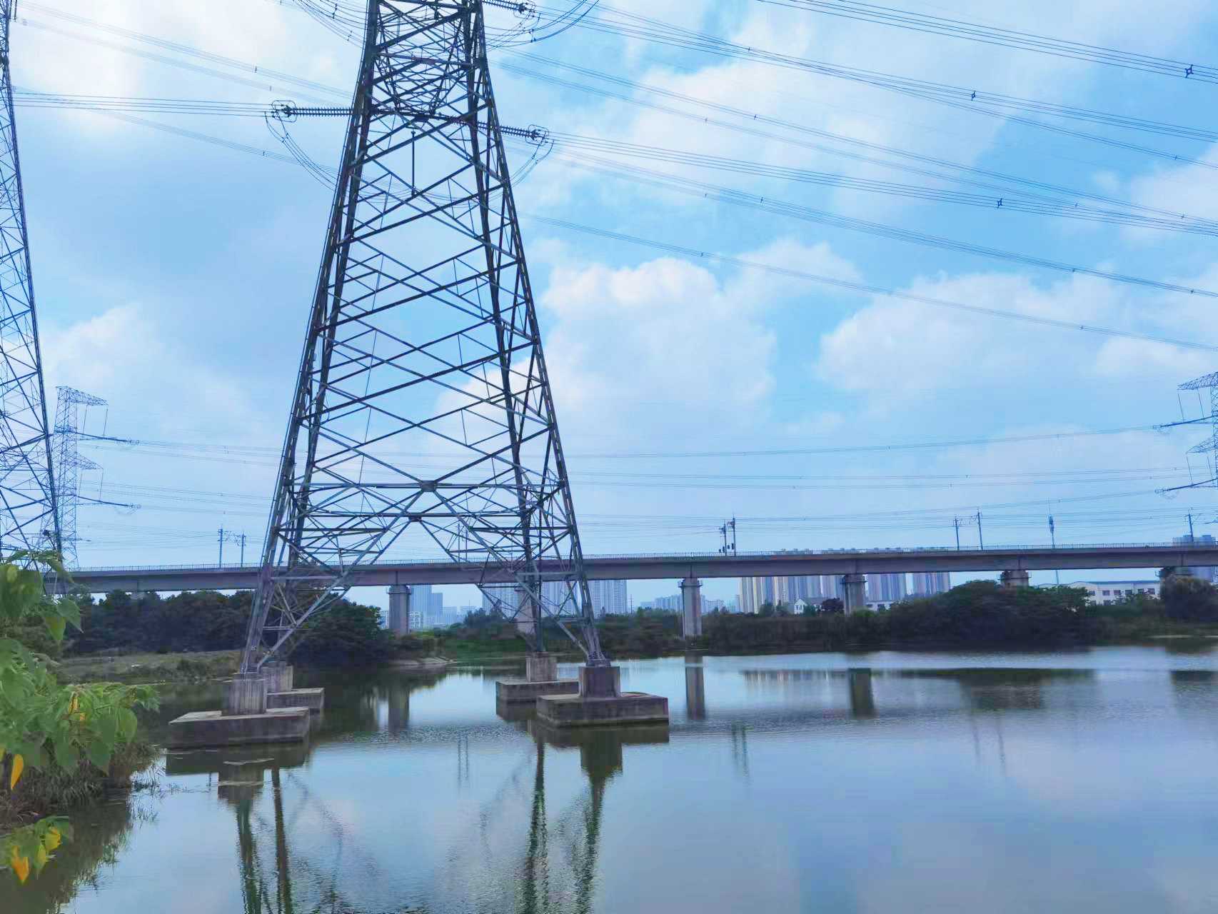 A tall steel electricity transmission tower stands beside a river, with a road bridge, distant city skyline, and power lines visible under a soft, warm-toned sky.
