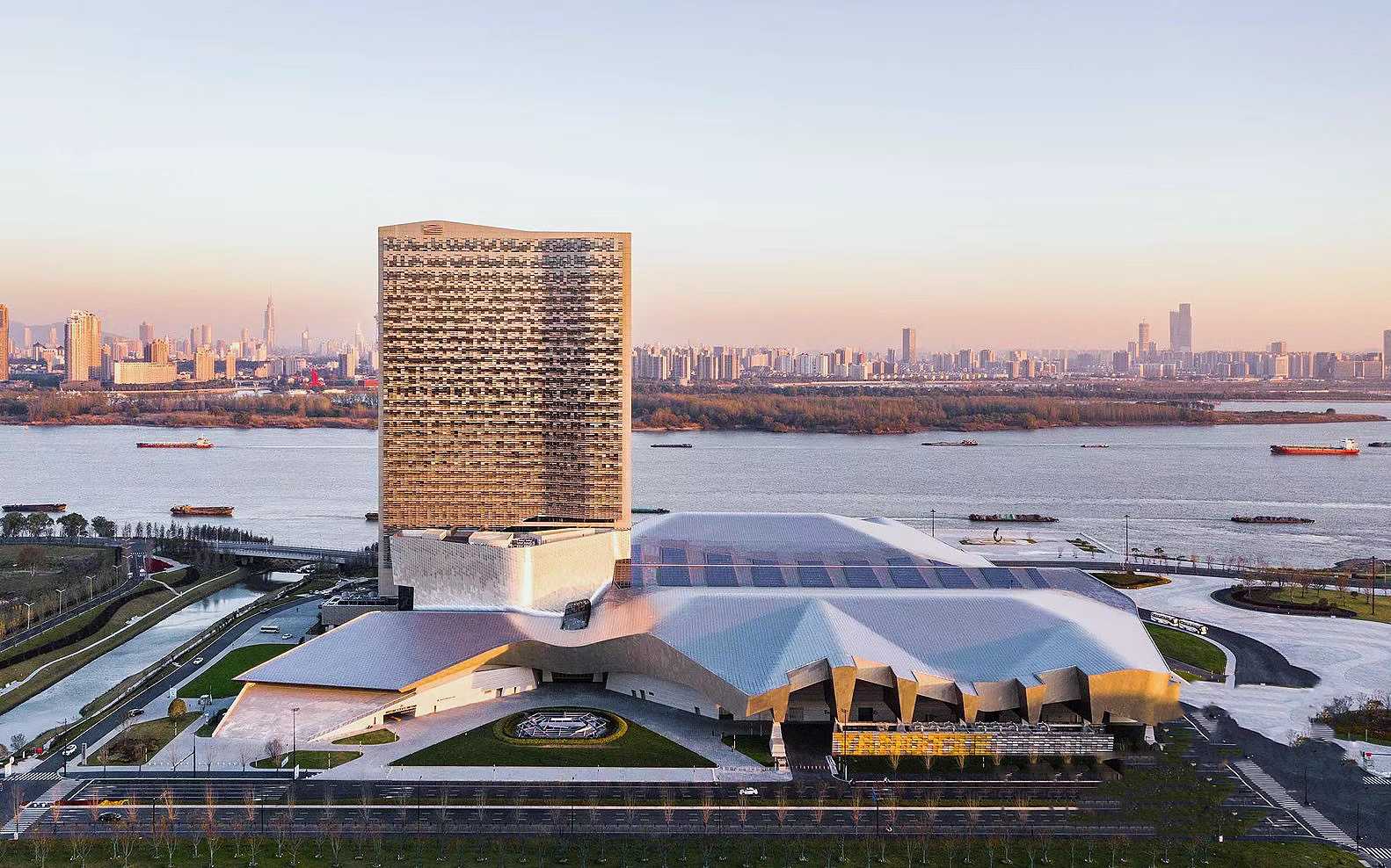 A wide aerial photograph at dusk showing a large modern building complex beside a calm river, with a tall patterned tower and wave-shaped roof glowing softly against the twilight sky.